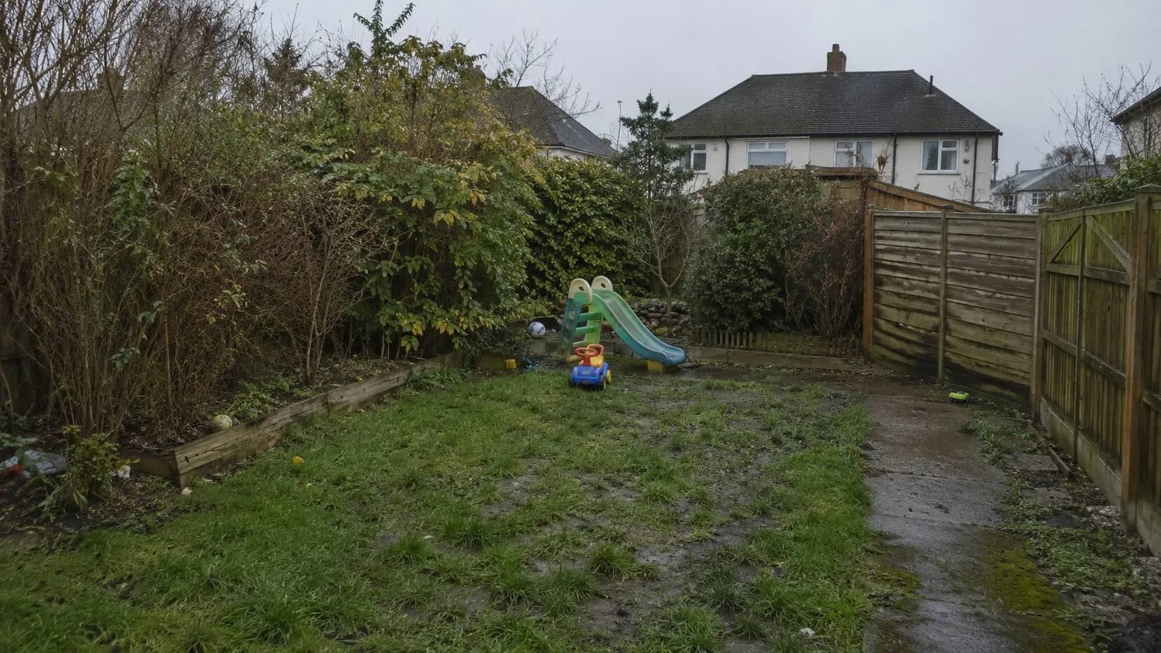 A tired suburban back garden before work: patchy worn lawn, a sagging rotten fence panel, an old plastic slide and an overgrown border.