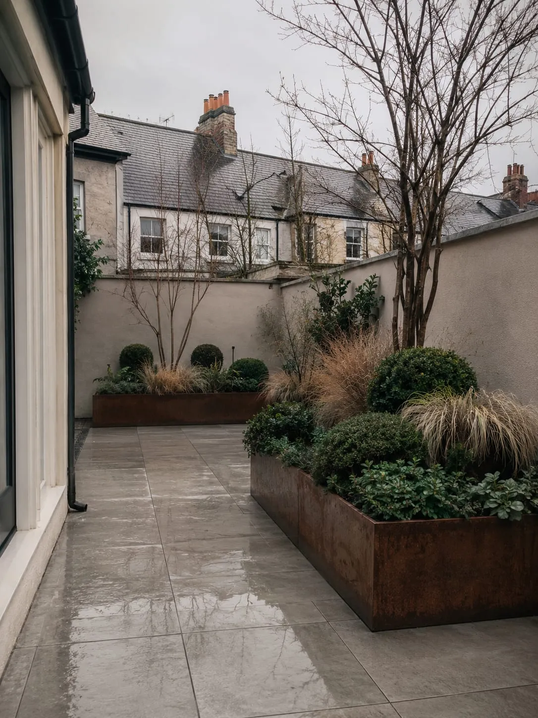 The same porcelain courtyard in winter, paving wet with rain and the corten planters holding clipped evergreen structure and cut-back grasses.